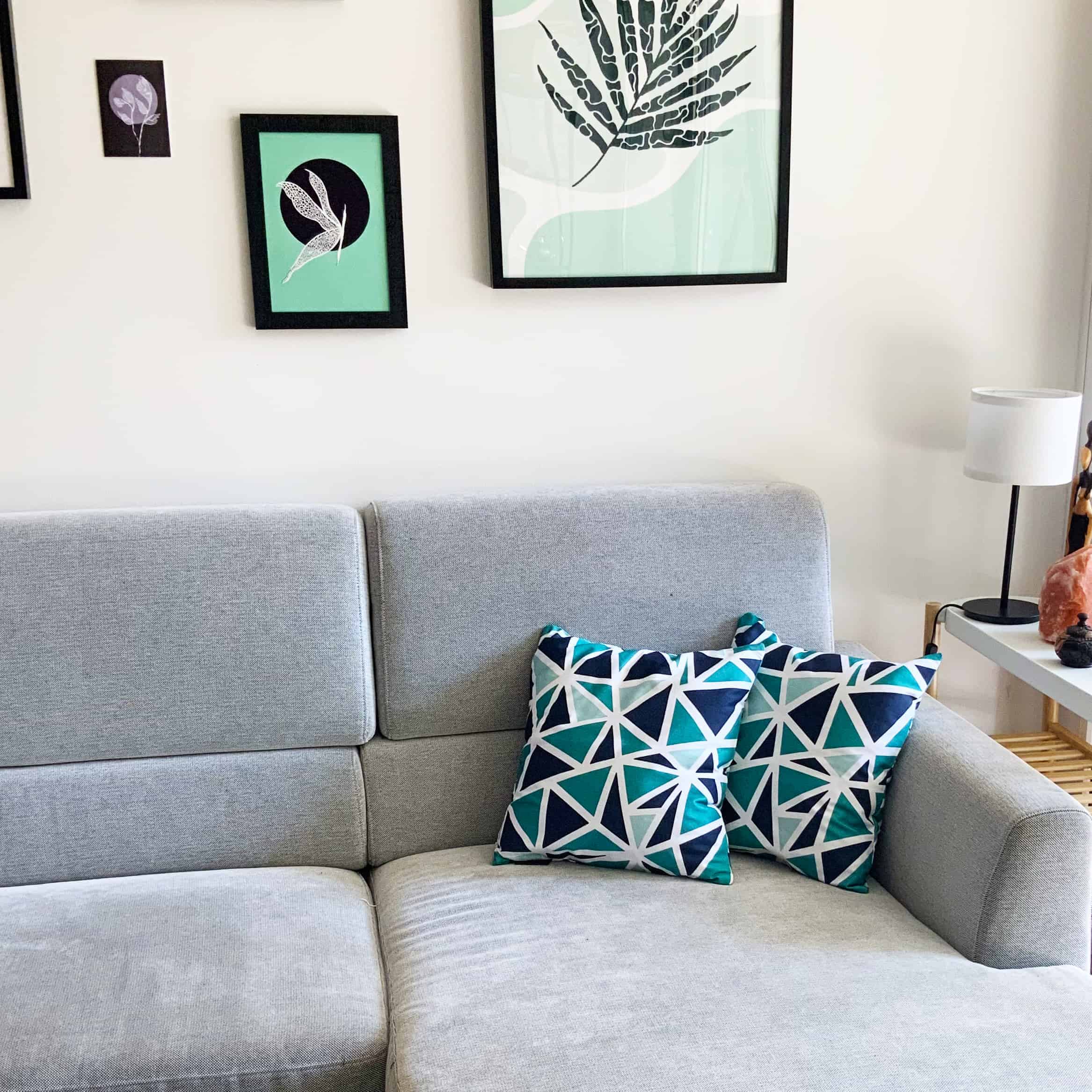Grey couch with two blue bold pattern cushions, some framed green artwork on the wall behind